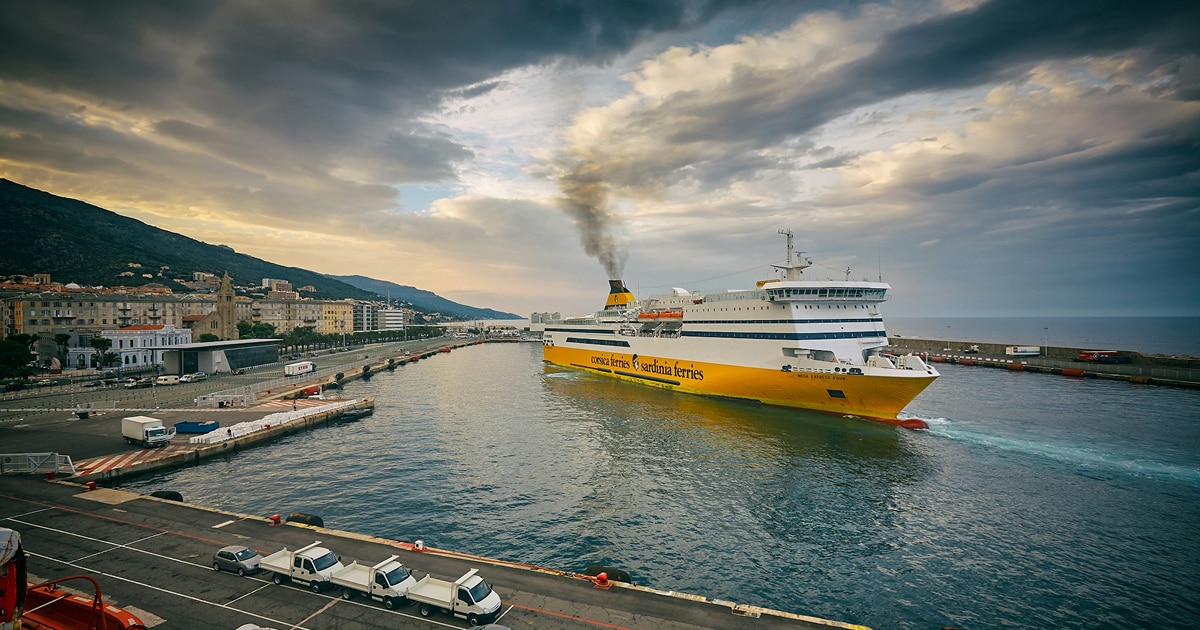 A scenic view of a yellow ferry departing from a harbor, emitting smoke, with mountains and a coastal town in the background.